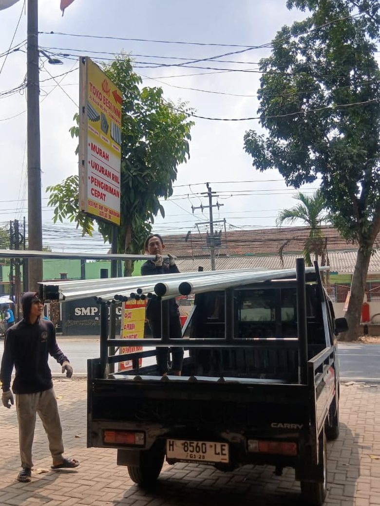 Dua orang staf Toko Besi INNI memuat baja ringan ke pikap di depan toko, dengan papan nama bertuliskan murah, ukuran riil, pengiriman cepat